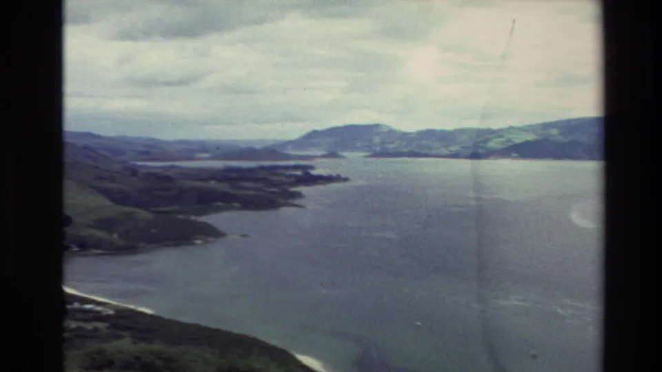 1982 New Zealand home-movie frame of a lake inlet and surrounding hills, seen through faded 8mm film with gate edges visible.