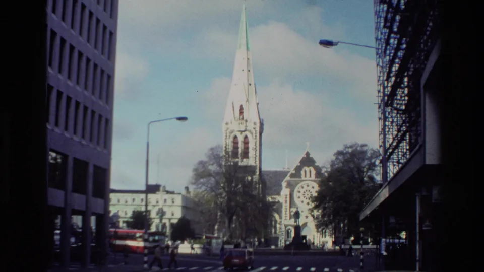 Christchurch Cathedral appears at the end of a city street, with pedestrians, cars, streetlights, trees, and tall commercial buildings framing the view.