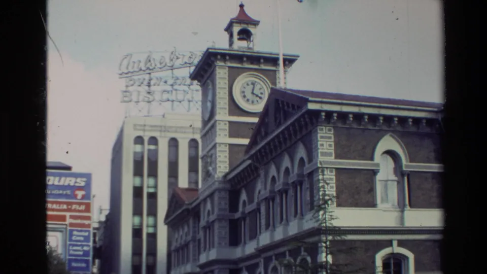 A Christchurch street frame shows a brick building with a clock tower beside taller commercial buildings and partially readable signage in the background.