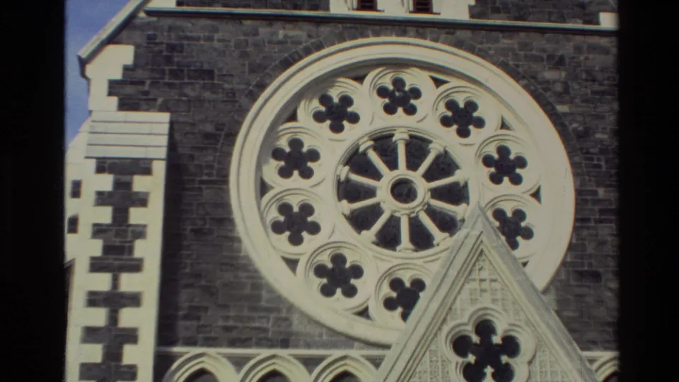 1982 New Zealand 8mm frame focused on a church rose window, dark stone wall, pale masonry trim and lower gable detail.
