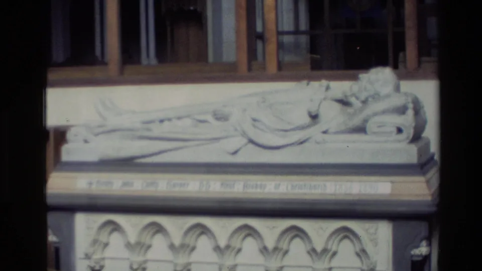 Visitor-shot 1982 New Zealand 8mm frame of a reclining stone effigy in a church interior, with carved base arches and an inscription band.