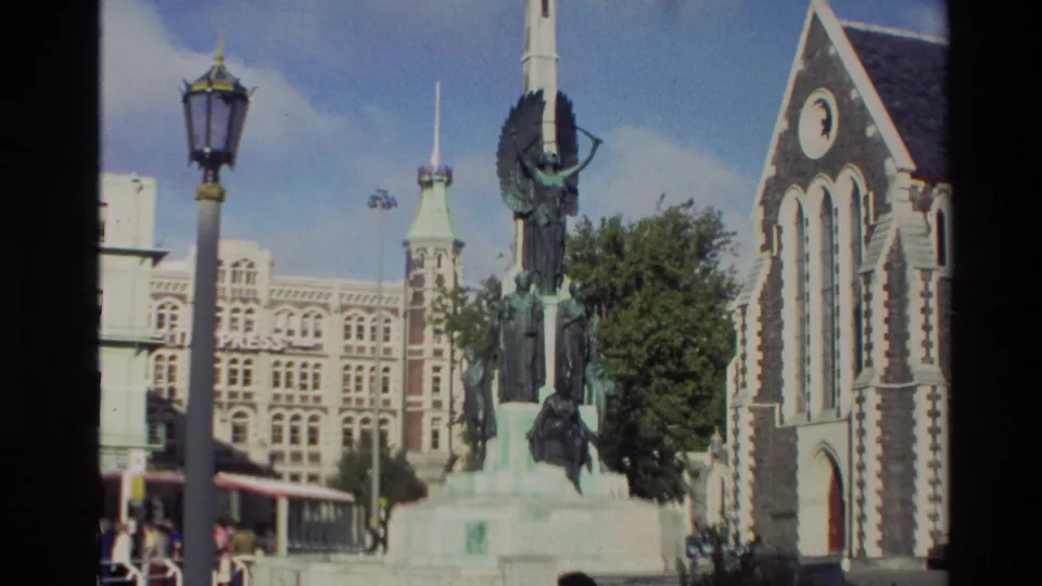 1982 New Zealand exterior frame showing a public memorial cross with sculptural figures beside historic buildings and a church facade.
