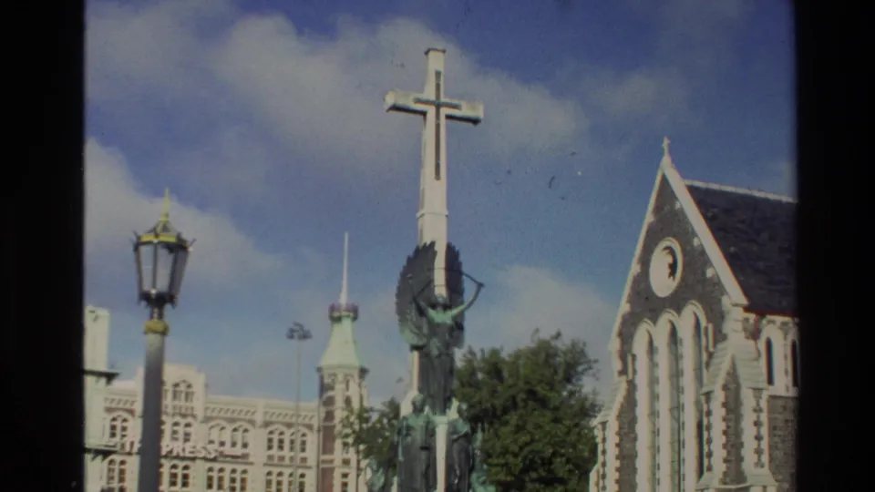 1982 New Zealand exterior frame showing the memorial cross and church facade, with historic buildings and trees behind them.