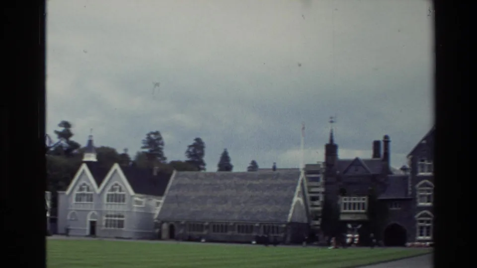 1982 New Zealand frame showing Christ's College lawn with gabled buildings, trees, and a towered stone structure at right.