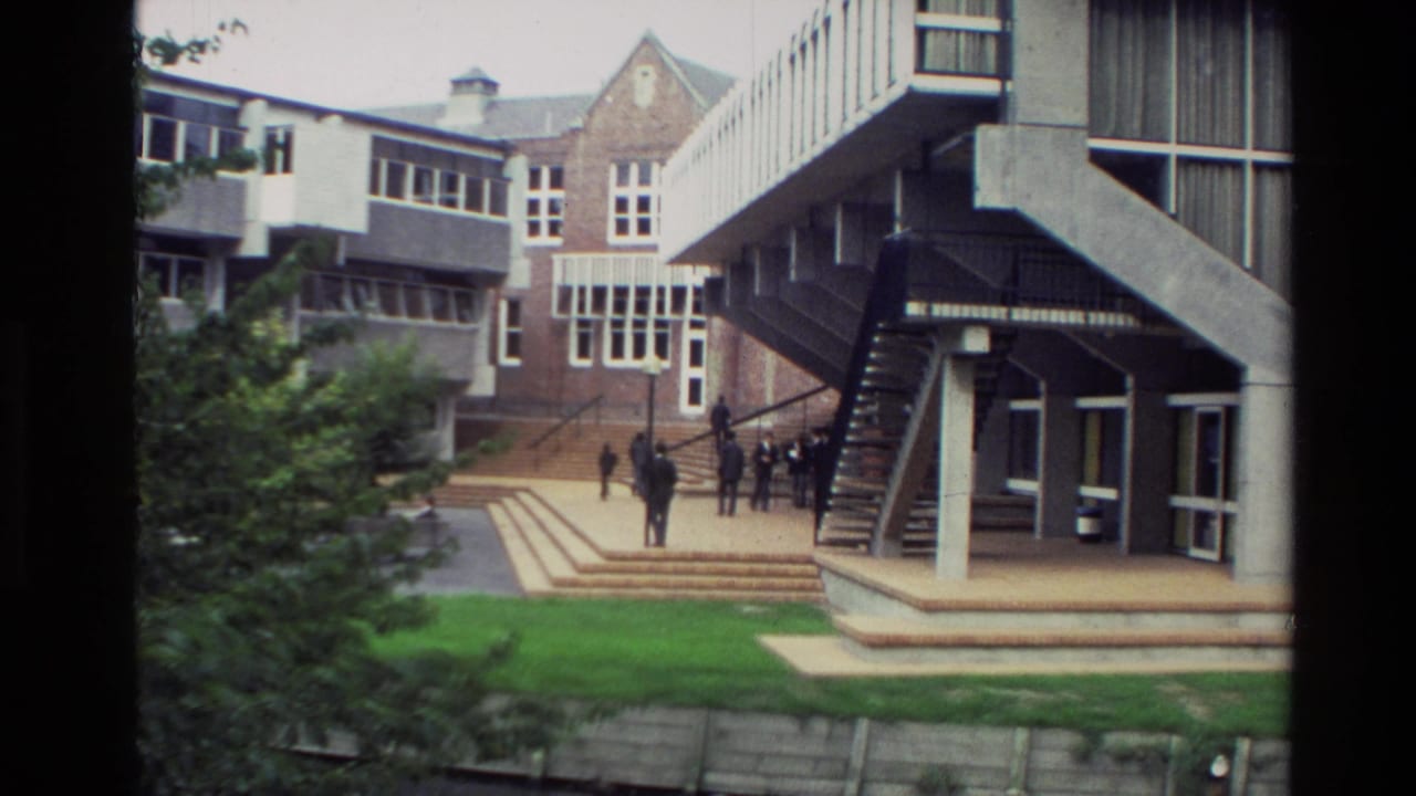 1982 New Zealand home-movie frame showing people near a modernist building complex, steps, lawn, and trees.