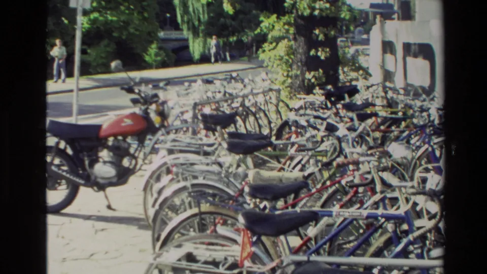 1982 New Zealand street-side scene with many parked bicycles, a red motorcycle, trees, and nearby pavement.