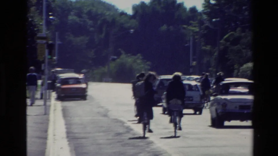 1982 New Zealand home-movie frame showing cyclists and compact cars on a tree-lined avenue.