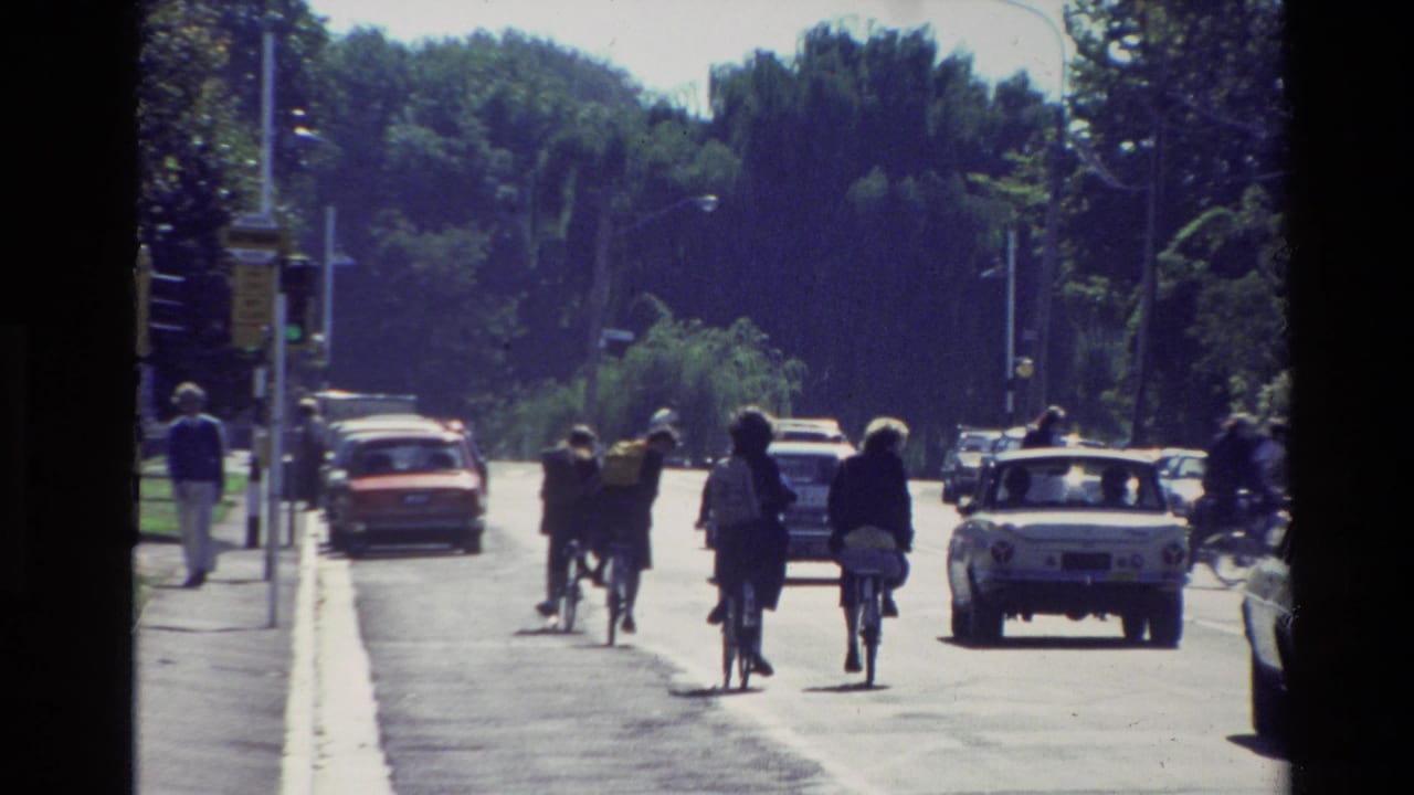 New Zealand 1982 street view showing people walking and cycling near traffic on a sunny day.