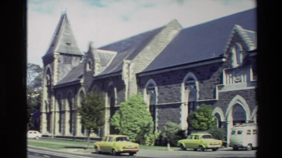 New Zealand 1982 street scene with a Gothic-style stone church, trees, parked cars, and a white van.