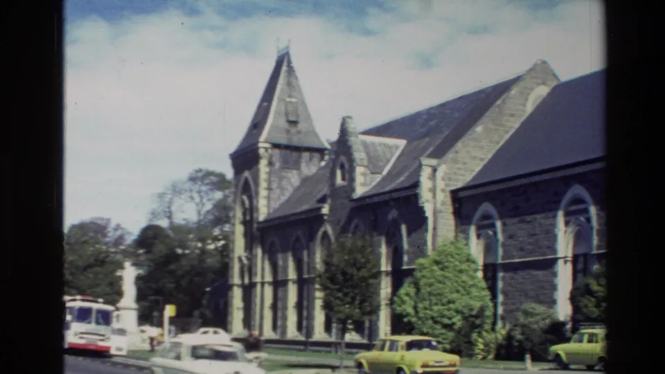 New Zealand 1982 view of a stone church facade with arched windows, trees, and cars at the roadside.