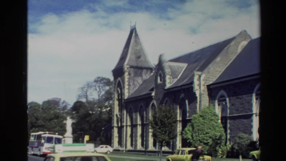 New Zealand 1982 street view of a stone church, leafy trees, parked vehicles, and bright sky.