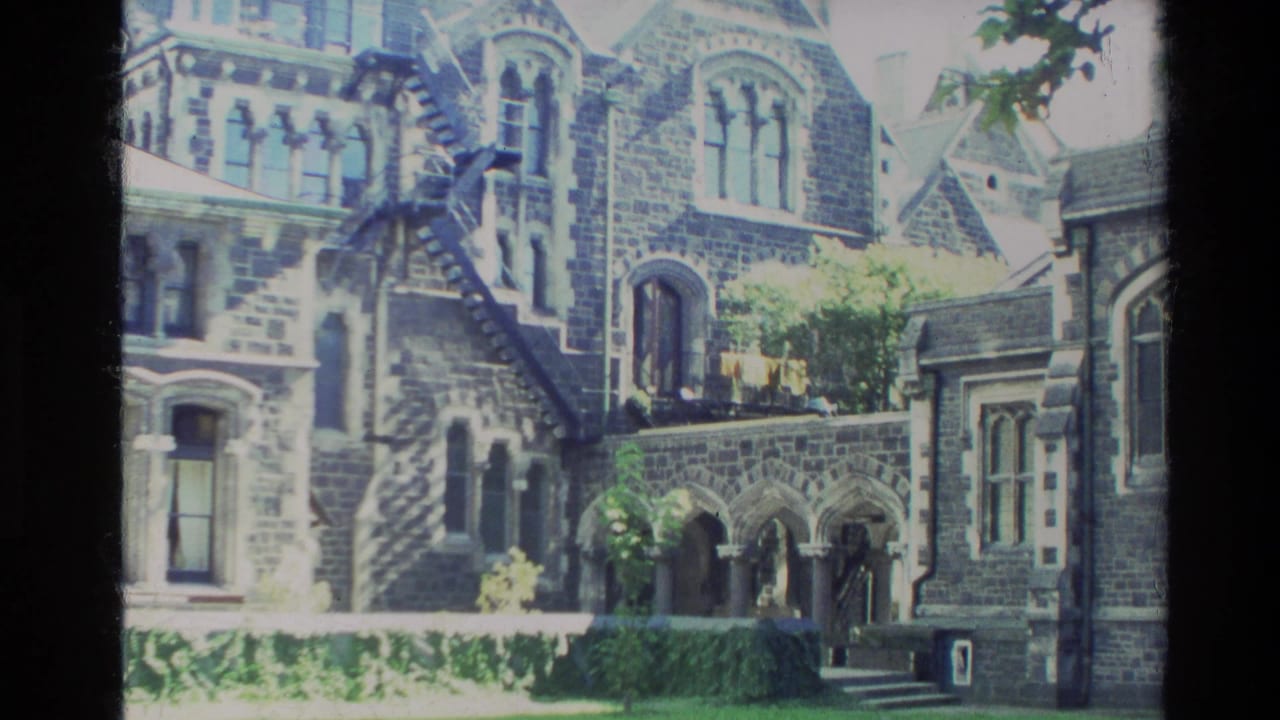 1982 New Zealand 8mm frame showing a stone church exterior, arched architectural details, courtyard greenery, and bright daylight exposure.
