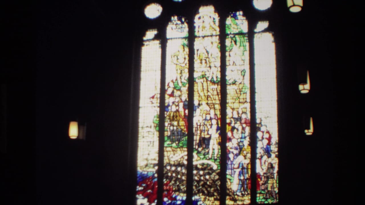 New Zealand 1982 home-movie frame showing a bright stained-glass window against a dark interior, with small hanging lights visible.