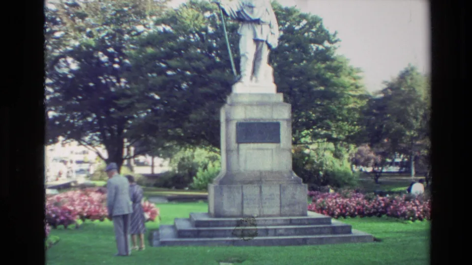 New Zealand 1982 home-movie frame of a robed stone statue with a staff on a pedestal, with two visitors standing near flowerbeds and trees.