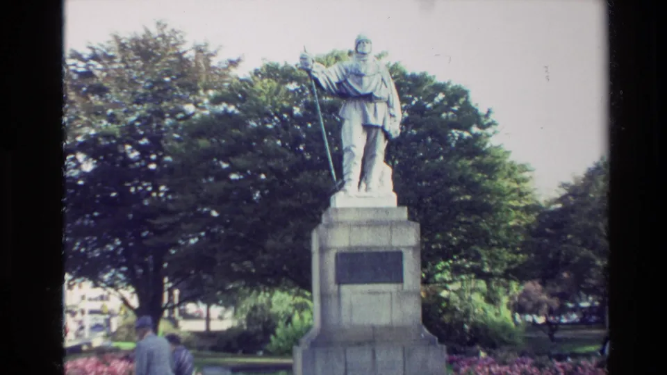 New Zealand 1982 home-movie frame with the robed statue centered on its pedestal, trees behind it, and a visitor partly visible in the foreground.