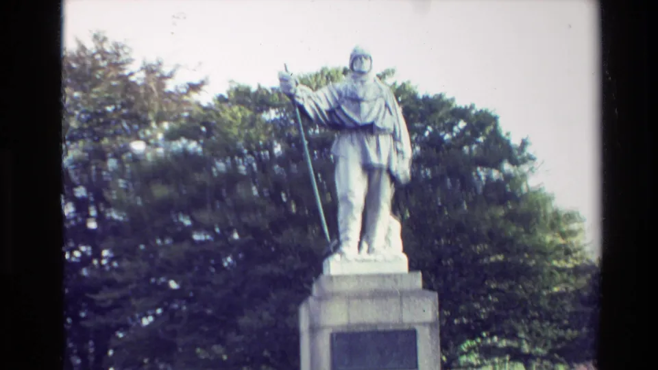New Zealand 1982 home-movie frame showing the upper pedestal and robed statue holding a staff, with dense trees behind it.