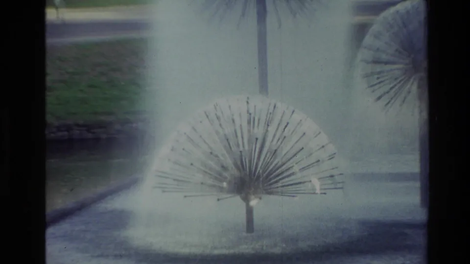 1982 New Zealand park footage showing a spherical fountain head spraying water near a curved pond edge, with lawn and palms behind it.