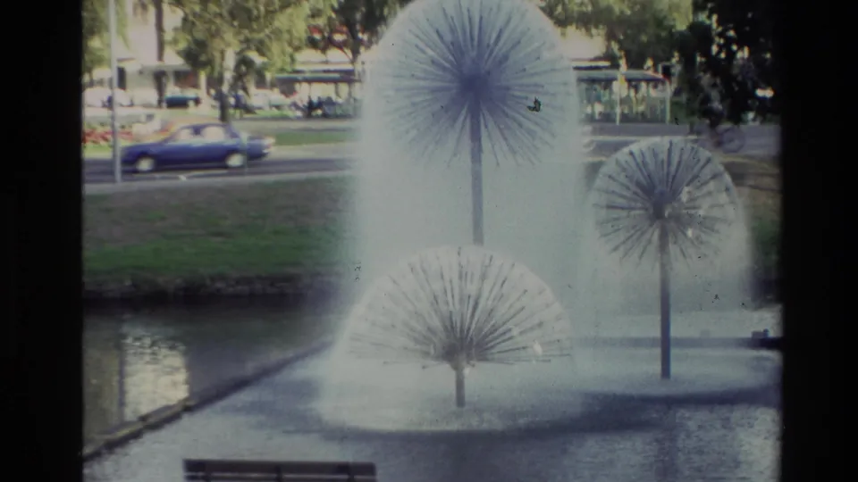 Wider 8mm view from 1982 New Zealand showing multiple dandelion-style fountains in a pond, with grass, trees, traffic, and a bench nearby.