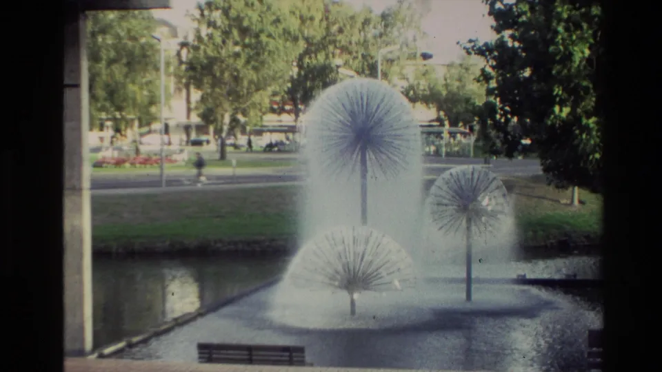1982 New Zealand 8mm frame showing several misting dandelion fountains in a park pond, with trees, a bench, lawn, and traffic in the distance.