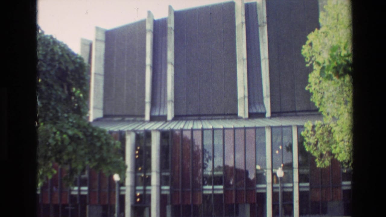 Modern building facade with tall concrete pillars, glass windows, trees, and lawn.