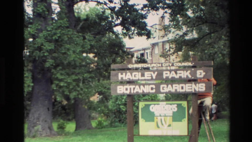 1982 New Zealand view of the Hagley Park and Botanic Gardens sign, with mature trees, a lawn, a building behind, and a person partly visible by a ladder.