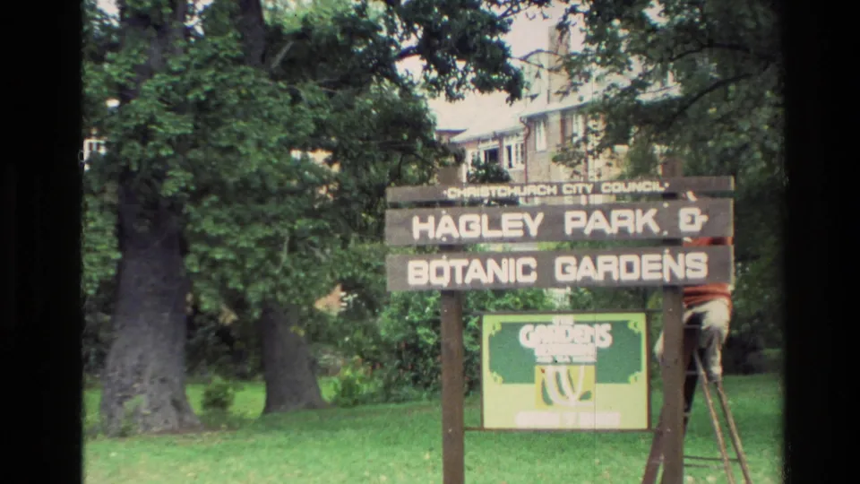 Closer 1982 New Zealand home-movie frame of the Hagley Park and Botanic Gardens sign, set on grass with large trees and a partly visible person on a ladder.
