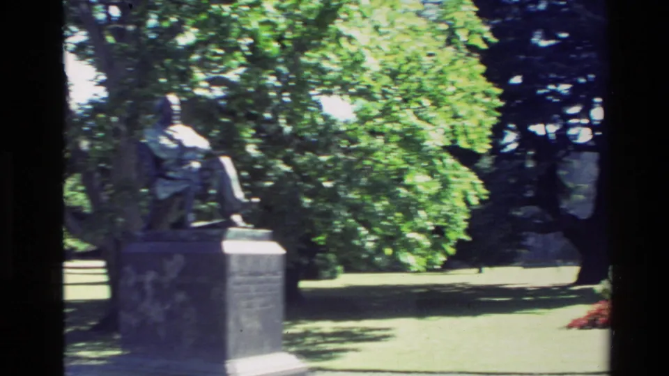 1982 New Zealand 8mm frame showing a stone statue on a pedestal near shaded lawn, mature trees, and red flowers.