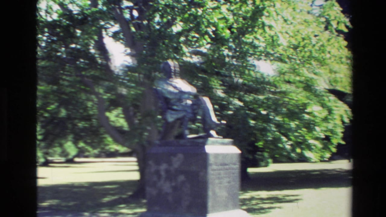 1982 New Zealand home-movie frame showing a seated bronze statue on a stone pedestal amid trees and lawn.