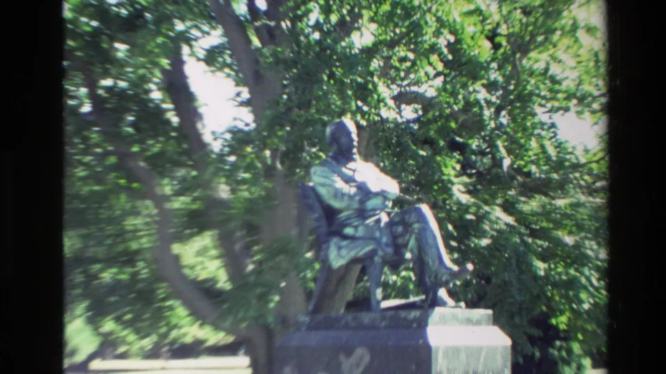 Closer handheld view of the seated bronze statue in a leafy New Zealand park, with strong sunlight and soft Super 8 detail.