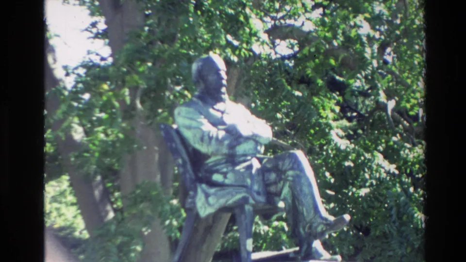 Close 8mm frame of the seated bronze figure, partly softened by focus and glare, with dense foliage behind it.