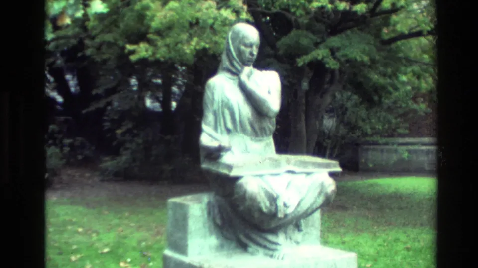 Soft 8mm view of a seated park statue in New Zealand, framed against trees and lawn with visible home-movie blur.