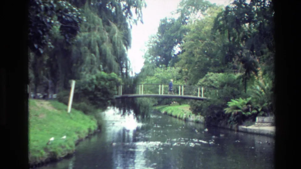 1982 New Zealand park scene with a narrow canal, arched footbridge, dense trees, and a distant person walking across.