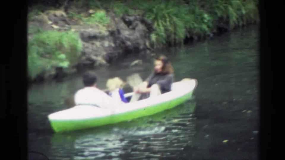 Close 8mm view of people paddling or riding in a small green-and-white boat on a New Zealand river in 1982.