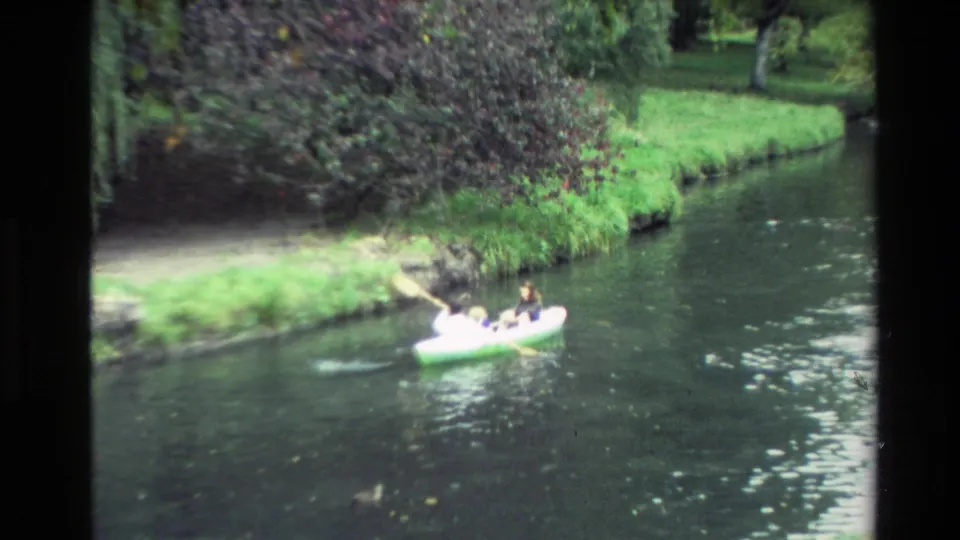 1982 New Zealand home-movie frame of a small boat carrying people along a calm river near grass and trees.