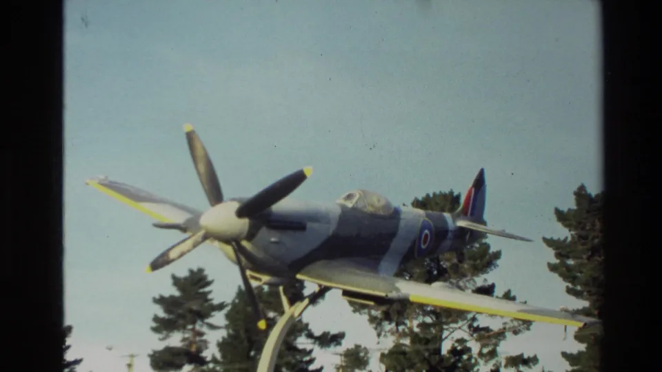 1982 New Zealand home-movie frame showing a mounted wartime-style propeller aircraft with roundel markings above trees.