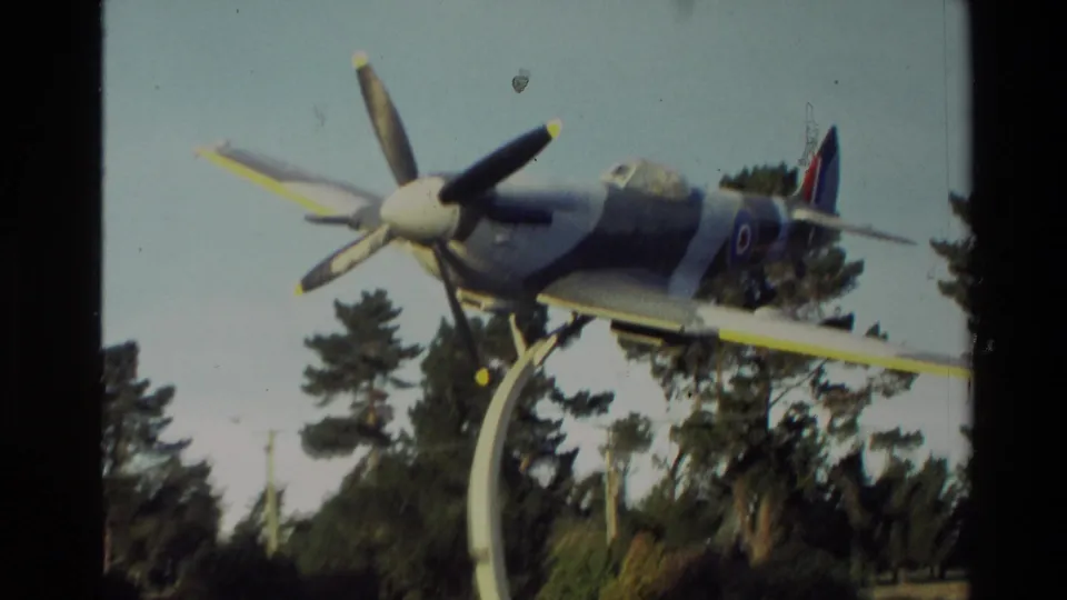 1982 New Zealand home-movie frame showing the aircraft display on a curved support, with pine trees and sky in the background.