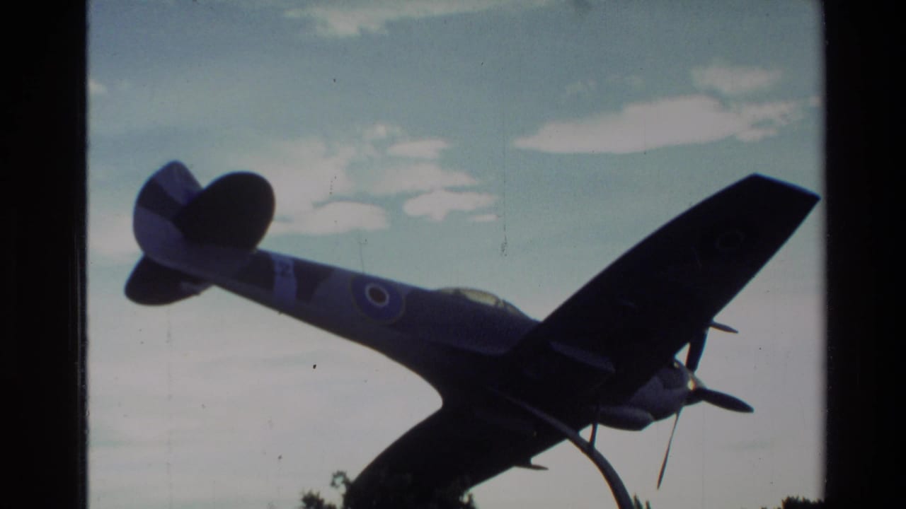 1982 New Zealand 8mm frame showing a dark blue fighter plane on display in silhouette against a pale cloudy sky.