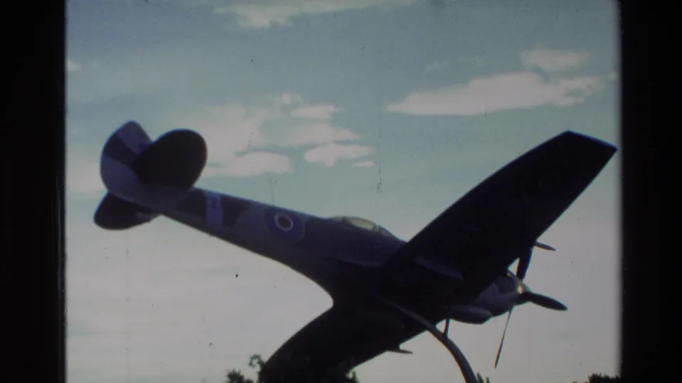 1982 New Zealand 8mm frame showing a dark blue fighter plane on display in silhouette against a pale cloudy sky.