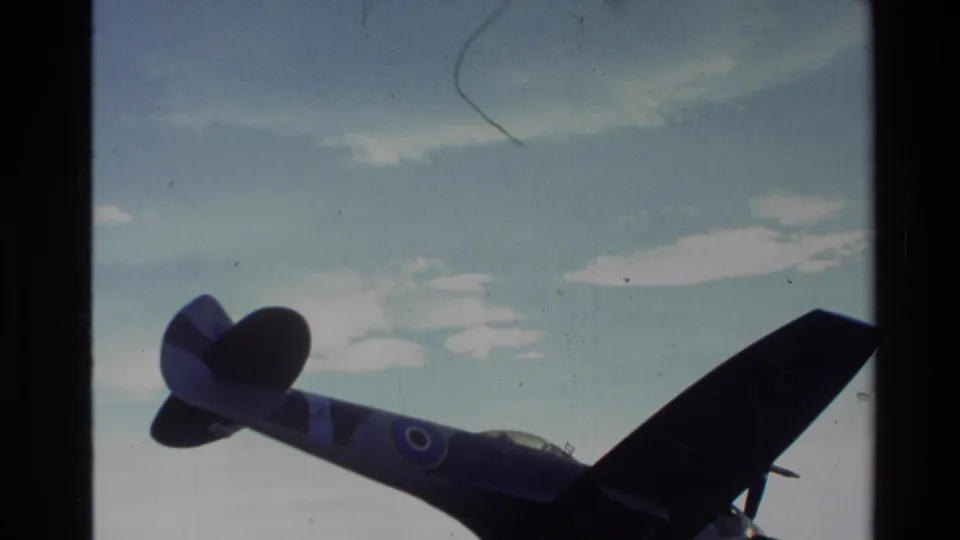 Archival 8mm view of a mounted fighter plane in New Zealand, with roundel markings faintly visible on the fuselage and wing.