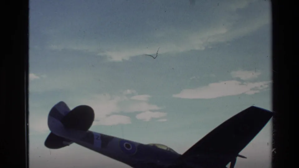 Home-movie frame of a fighter plane display, partly silhouetted with visible roundel markings and soft 8mm color fading.