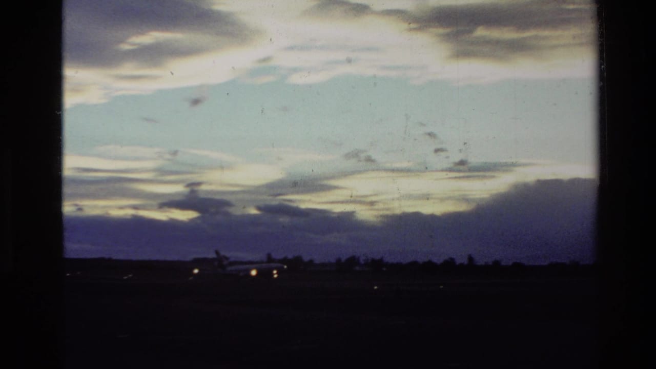1982 New Zealand 8mm frame showing a low airport horizon, glowing runway or aircraft lights, and a clouded dusk sky.