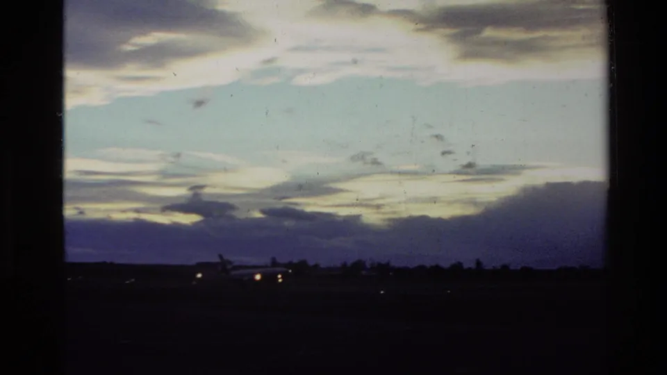 1982 New Zealand 8mm frame showing a low airport horizon, glowing runway or aircraft lights, and a clouded dusk sky.