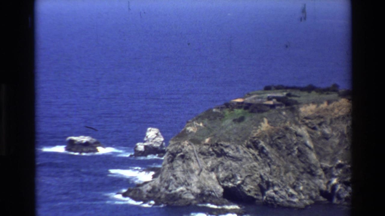1980 8mm view of the Big Sur coast, showing a rocky cliff edge, sea stacks, white surf, and deep blue Pacific water.