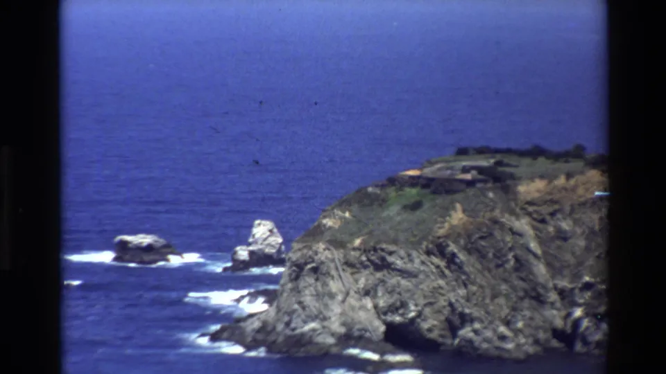 Big Sur coastline in 1980 home-movie footage, with sea stacks, a steep rocky point, white breaking waves, and open Pacific water.