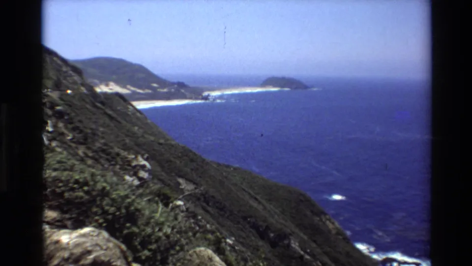 1980 8mm coastal overlook near Big Sur, showing a sloping foreground cliff, distant sandy shoreline, headland, and blue Pacific water.