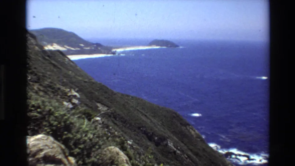 Big Sur coastal view from 1980 8mm footage, with a sloped cliff in the foreground, distant beach, white surf, and offshore blue water.