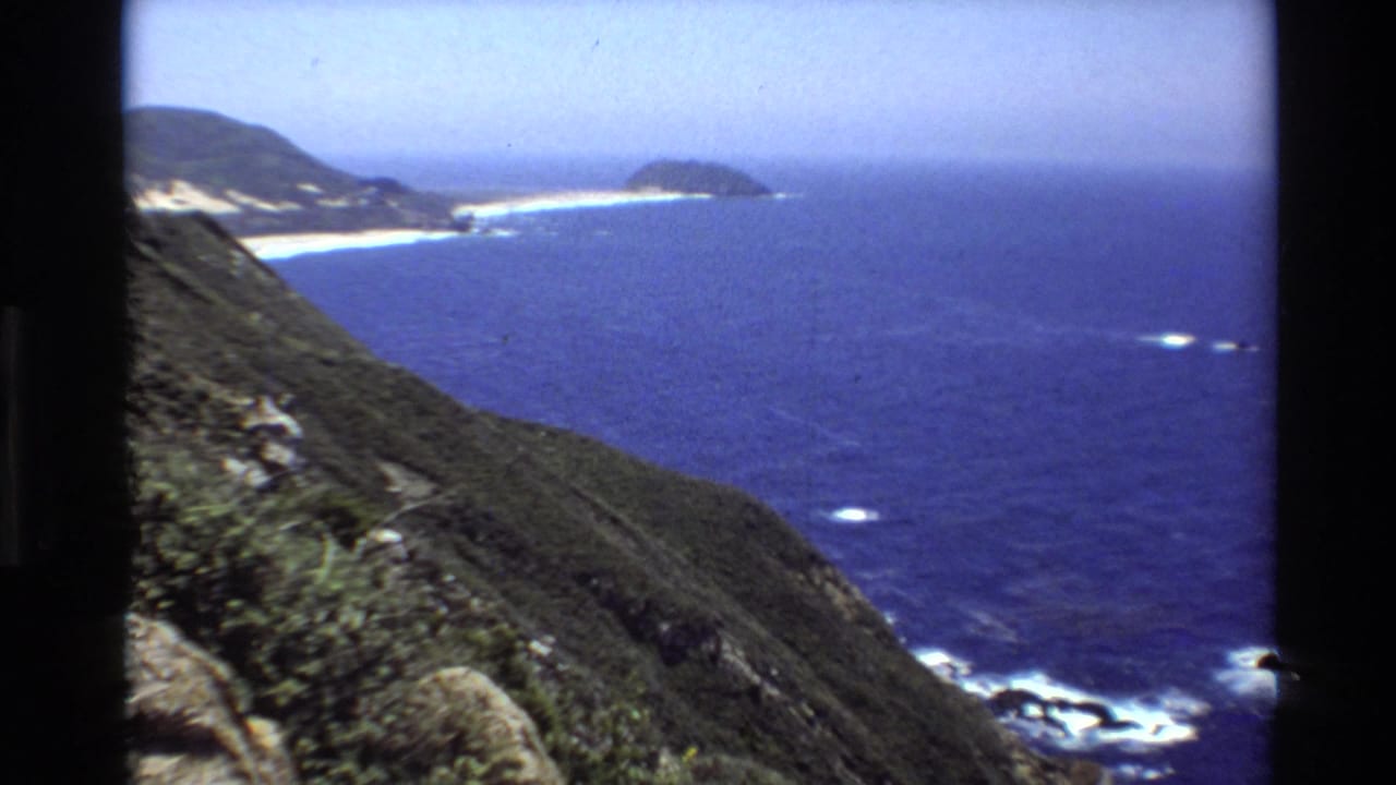 1980 Big Sur home-movie frame showing steep green cliffs above blue Pacific water, with a distant sandy beach and white surf.