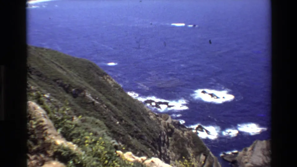 1980 Big Sur coastal frame showing a steep green hillside dropping toward rocks and foamy Pacific surf below.