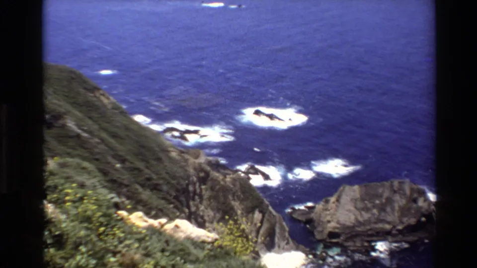 1980 Big Sur frame showing rocky shoreline, foamy Pacific waves, and steep green coastal slopes from an elevated viewpoint.