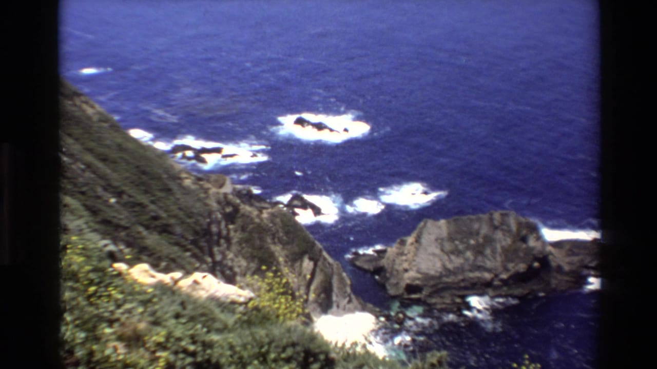 1980 8mm Big Sur cliffside view with deep blue Pacific water, breaking white surf, jagged rocks, and coastal vegetation in the foreground.